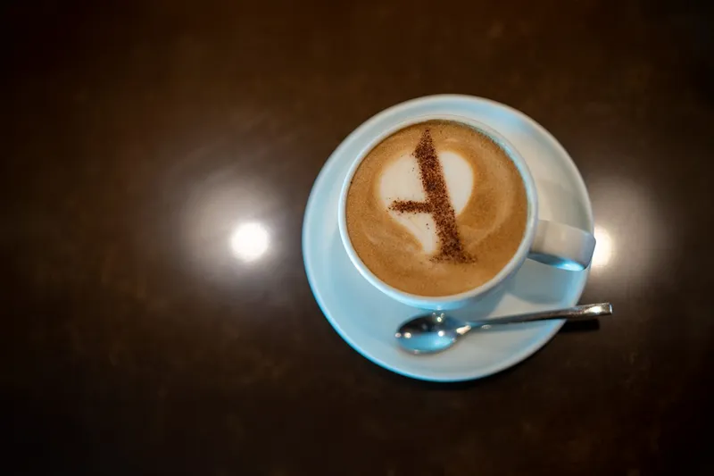 A latte with a letter "A" made of cocoa on the foam, served in a white cup and saucer with a spoon.