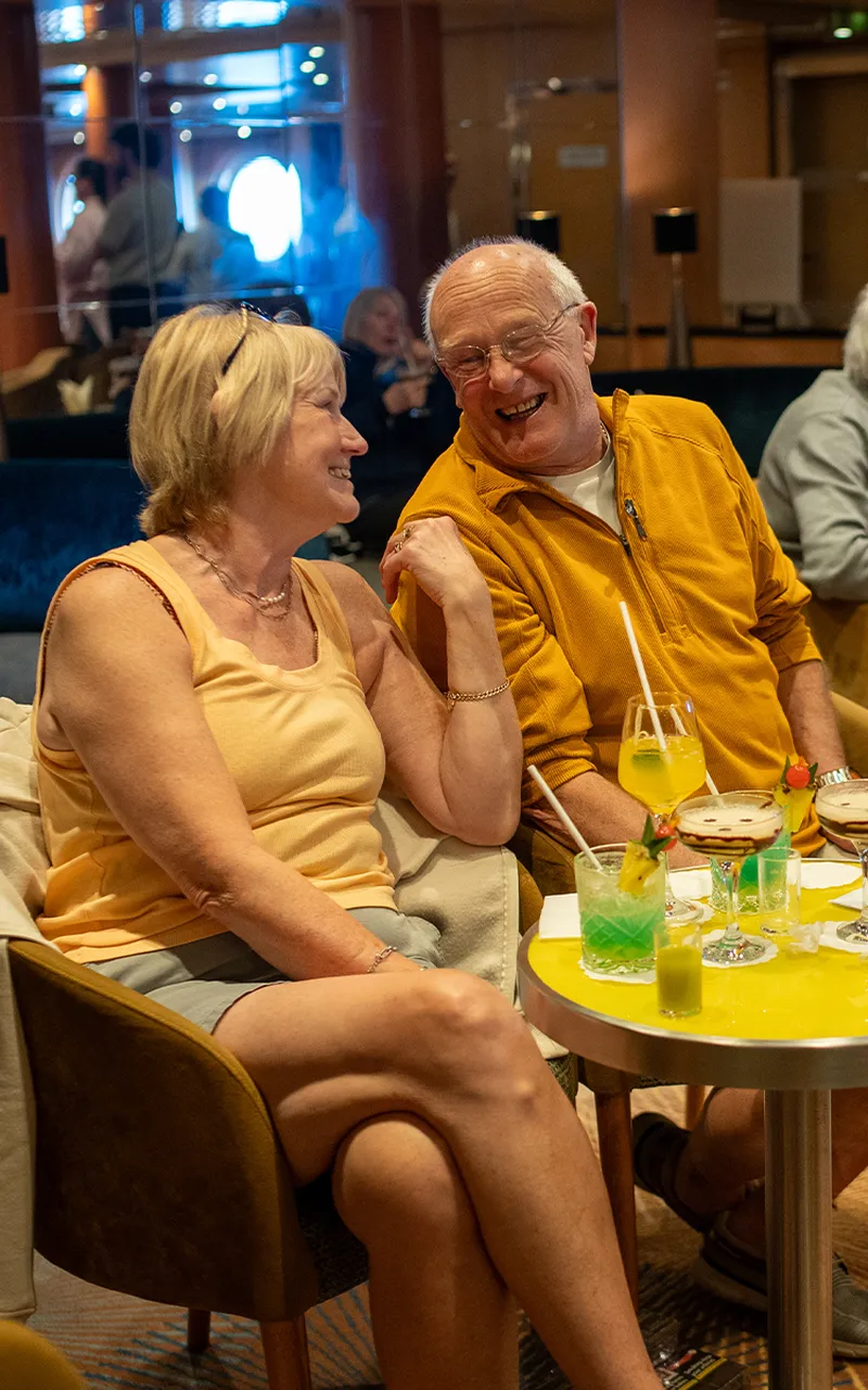 Two people sitting in a lounge, smiling at each other. Drinks are on the table, including cocktails with garnishes.