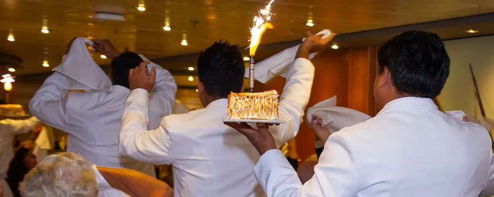 Waitstaff in white uniforms carry baked alaska with a sparkler through a restaurant