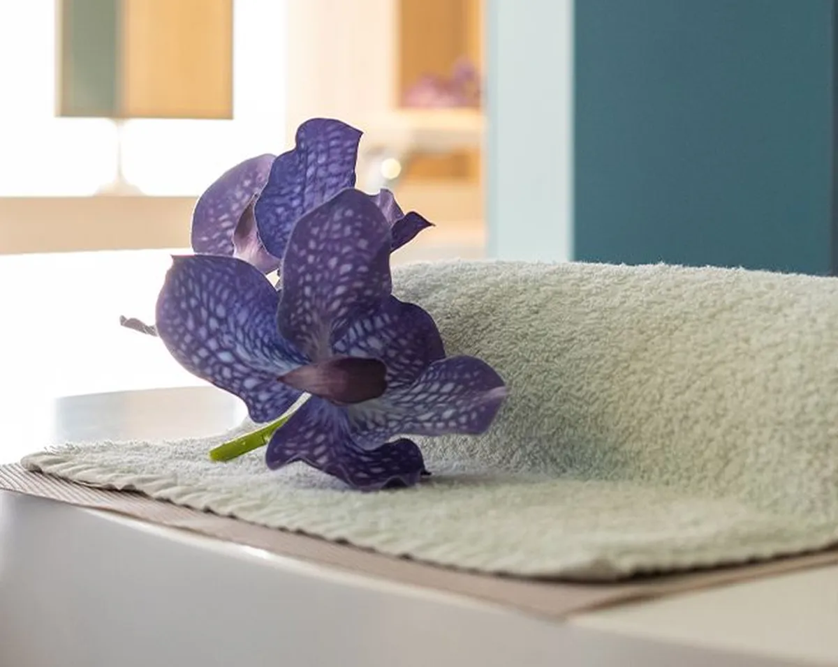 A purple flower rests on a folded towel atop a white surface, with a modern chair and soft lighting in the background.