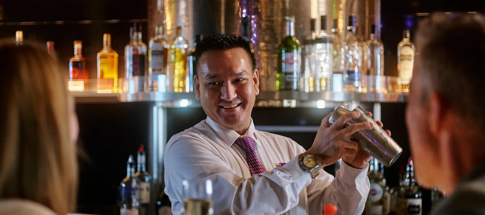 Bartender shaking a cocktail shaker, smiling at guest seated at a bar.