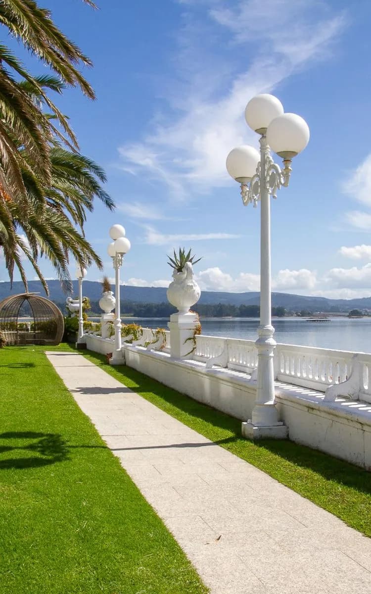 Pathway with white street lamps and palm trees beside a calm waterfront in Vigo, under a clear blue sky.