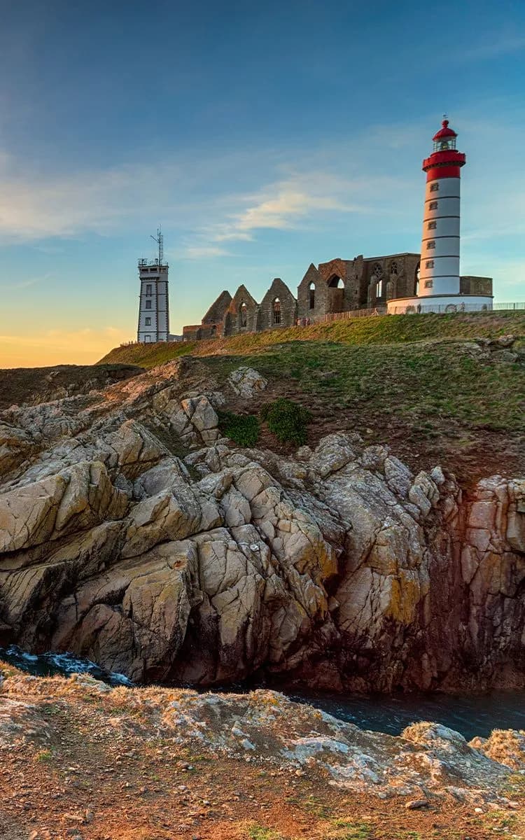 A rocky cliff with a red and white lighthouse and ruins at sunset in Brest, under a partly cloudy sky.