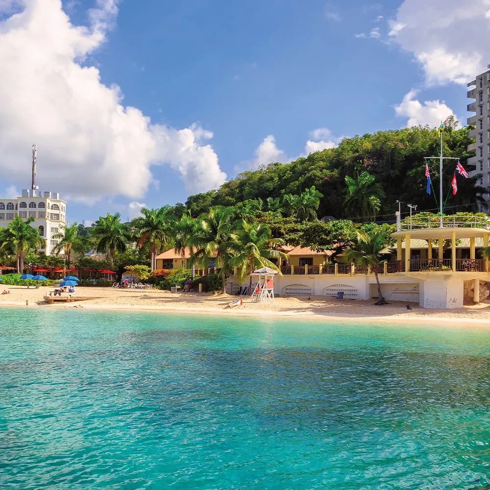 Beachfront scene with turquoise water, sandy shore, palm trees, and colorful buildings in Jamaica