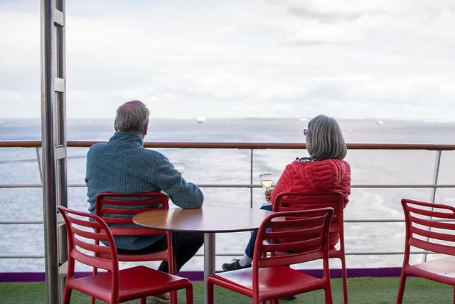 Two people sitting at a table on a ship's deck, looking out at the ocean, holding wine glasses.