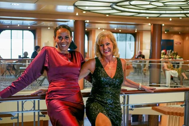 Kerrie Ann and Julie in elegant evening dresses pose together, smiling, on a balcony inside a stylish, well-lit venue.