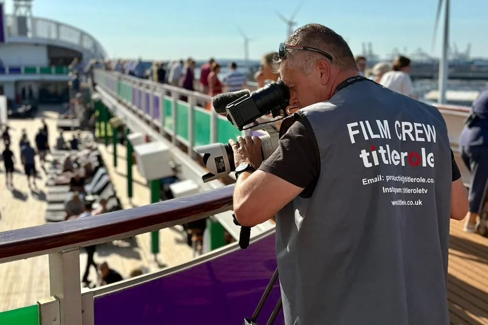 Film crew member with a camera filming on a ship's deck, wearing a vest labeled "FILM CREW titlerole." Passengers and deck chairs are visible.
