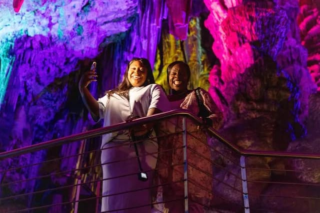 Jacque & Tamara smile on a railing inside a cave, illuminated by colorful lights in pink, purple, and yellow hues.