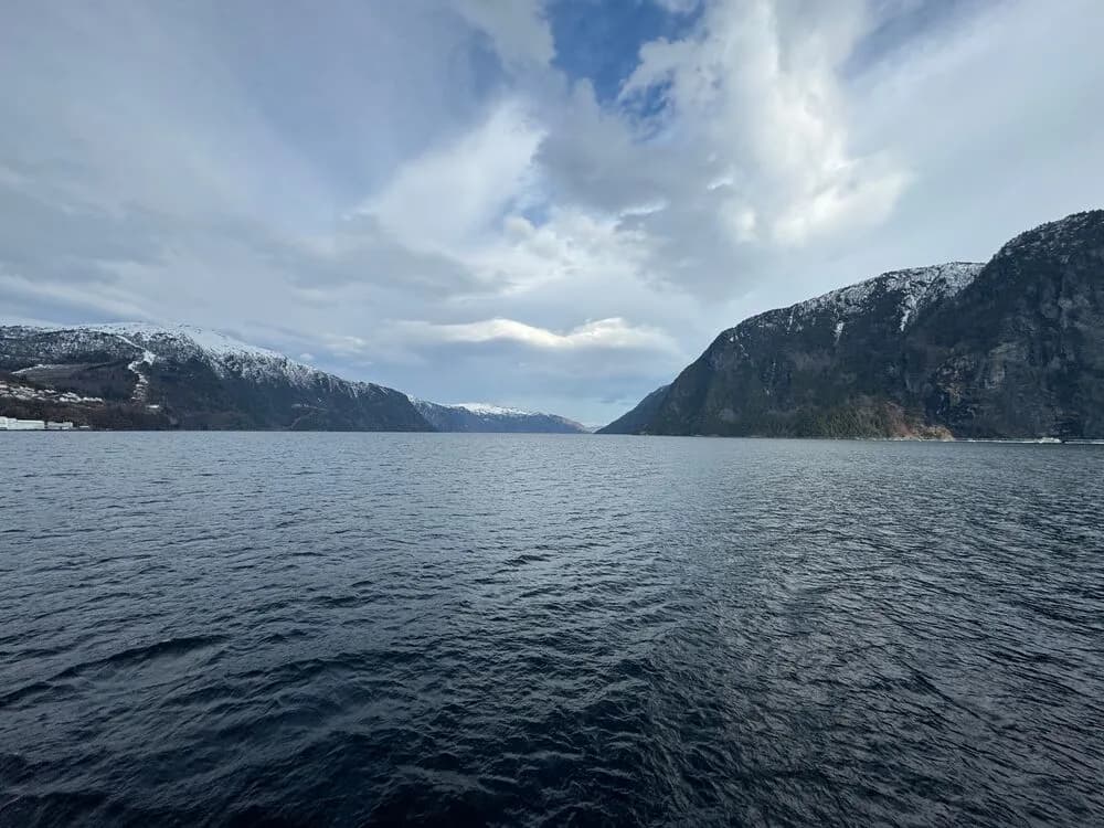 A serene fjord with calm water, surrounded by snow-capped mountains under a partly cloudy sky.