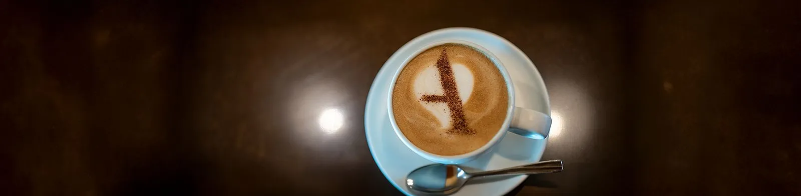 A latte with a letter "A" made of cocoa on the foam, served in a white cup and saucer with a spoon.