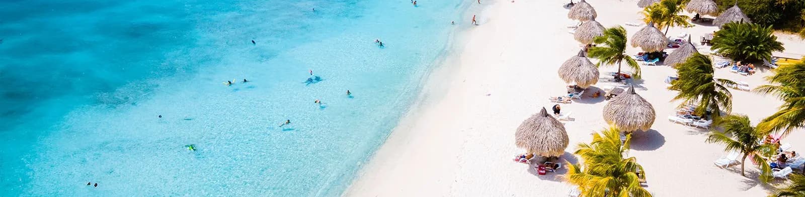 Aerial view of a beach in Curacao with turquoise water, white sand, people swimming, and straw umbrellas surrounded by palm trees.