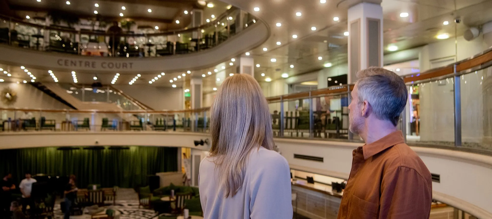 A man and woman overlook the spacious, Centre Court , surrounded by seating, and lit with numerous ceiling lights.