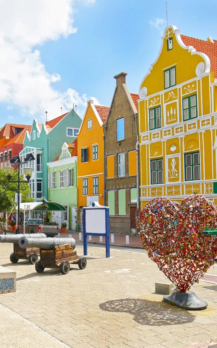 Colorful buildings with a heart-shaped sculpture covered in locks and an old cannon on a sunny day.