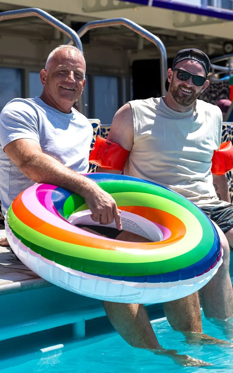 Graham & Lewis sitting by a pool; one holds a rainbow inflatable ring, both smiling. One wears water wings and sunglasses.