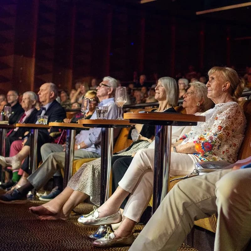 Audience seated in the Palladium theater, attentively watching a performance. Tables with drinks are in front of them, and the setting is dimly lit.