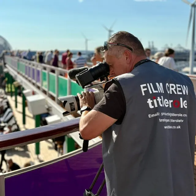 Film crew member with a camera filming on a ship's deck, wearing a vest labeled "FILM CREW titlerole." Passengers and deck chairs are visible.