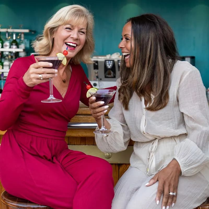 Kerrie Ann and Julie laughing and holding cocktails at a bar. One wears a red outfit, the other white. The background features shelves with bottles.