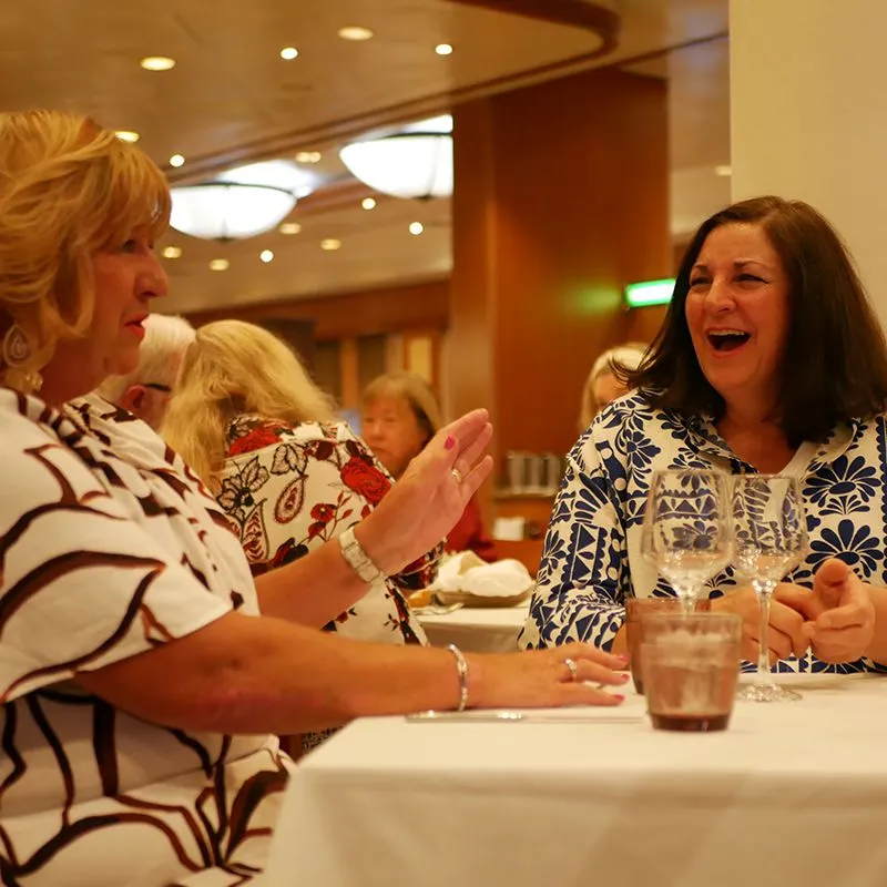Anne & Yvonne laughing and talking at a restaurant table, surrounded by other diners. The setting is warmly lit and lively.